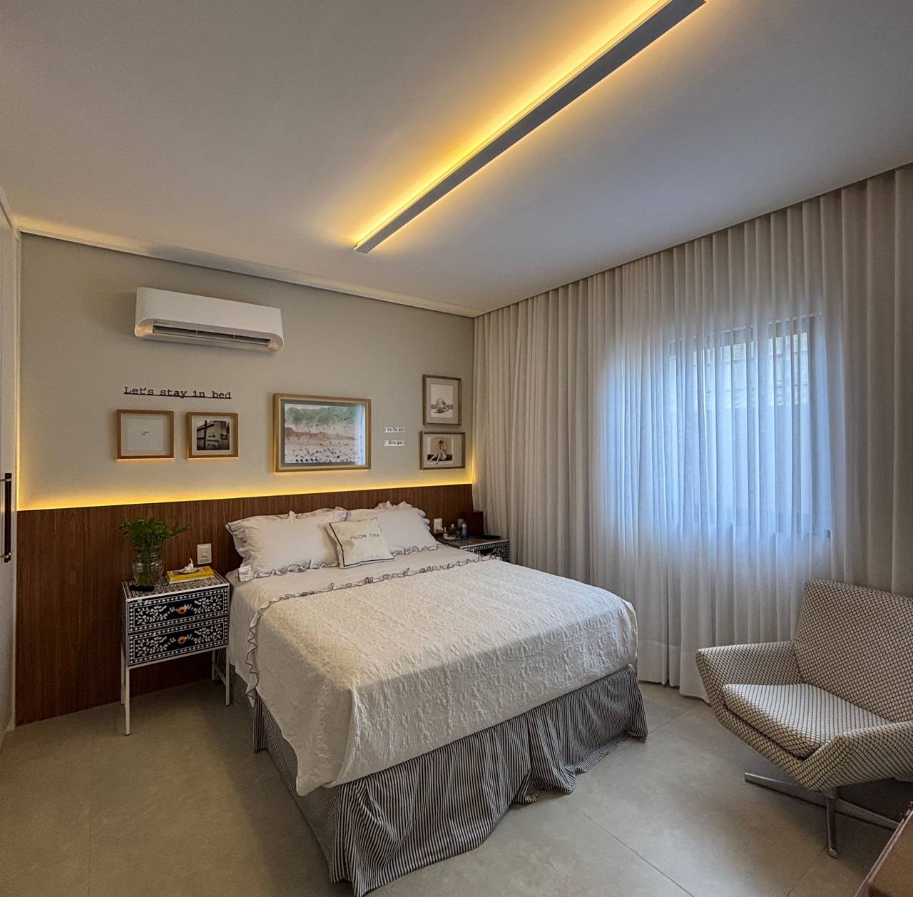 Quarto com cama clara, iluminação indireta, painel amadeirado, cortina e poltrona